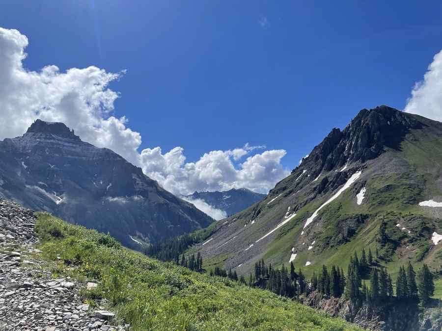 A green alpine basin between two dark peaks, snowfields on the slopes and white clouds overhead