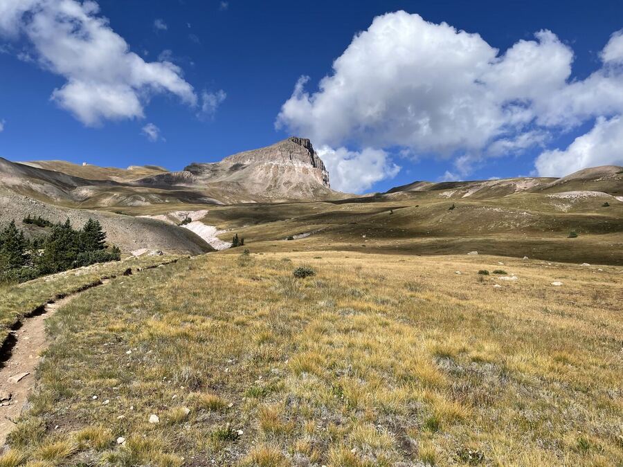 A grassy bench and a trail leading toward a blocky summit under big clouds