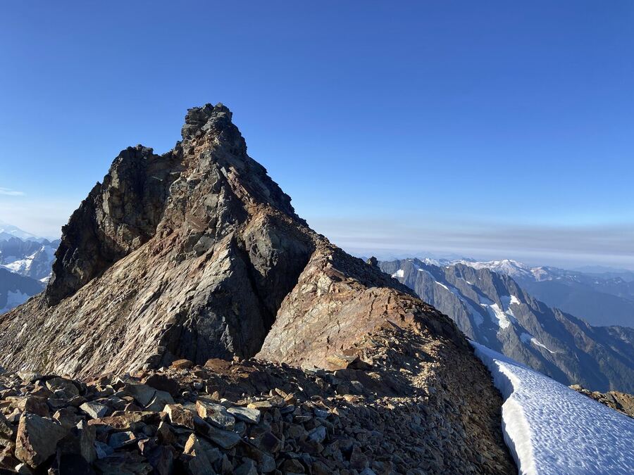 A sharp rock summit, a snowfield, and a range beyond