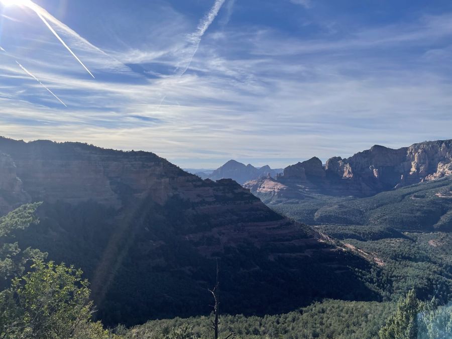 Red rock cliffs and a forested canyon in hazy afternoon light, contrails across the sky