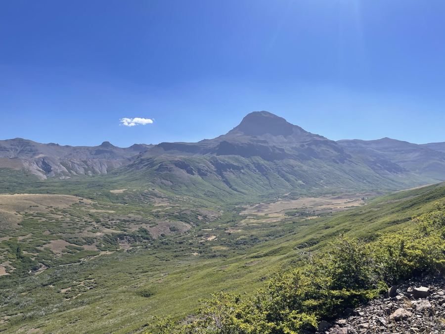 A broad green valley in late light below a single rounded peak, a small lake in the distance